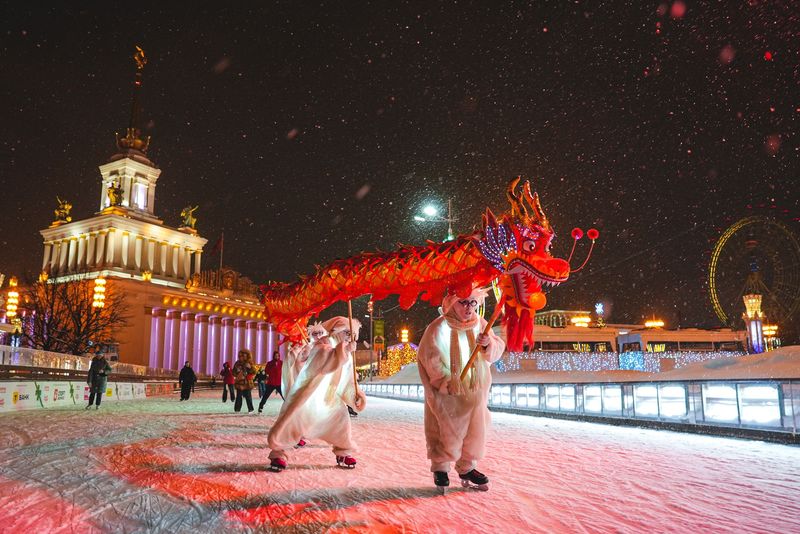Ночной каток на ВДНХ в снегопад: люди в костюмах несут красного китайского дракона по льду, вокруг подсвеченные павильоны и колесо обозрения.