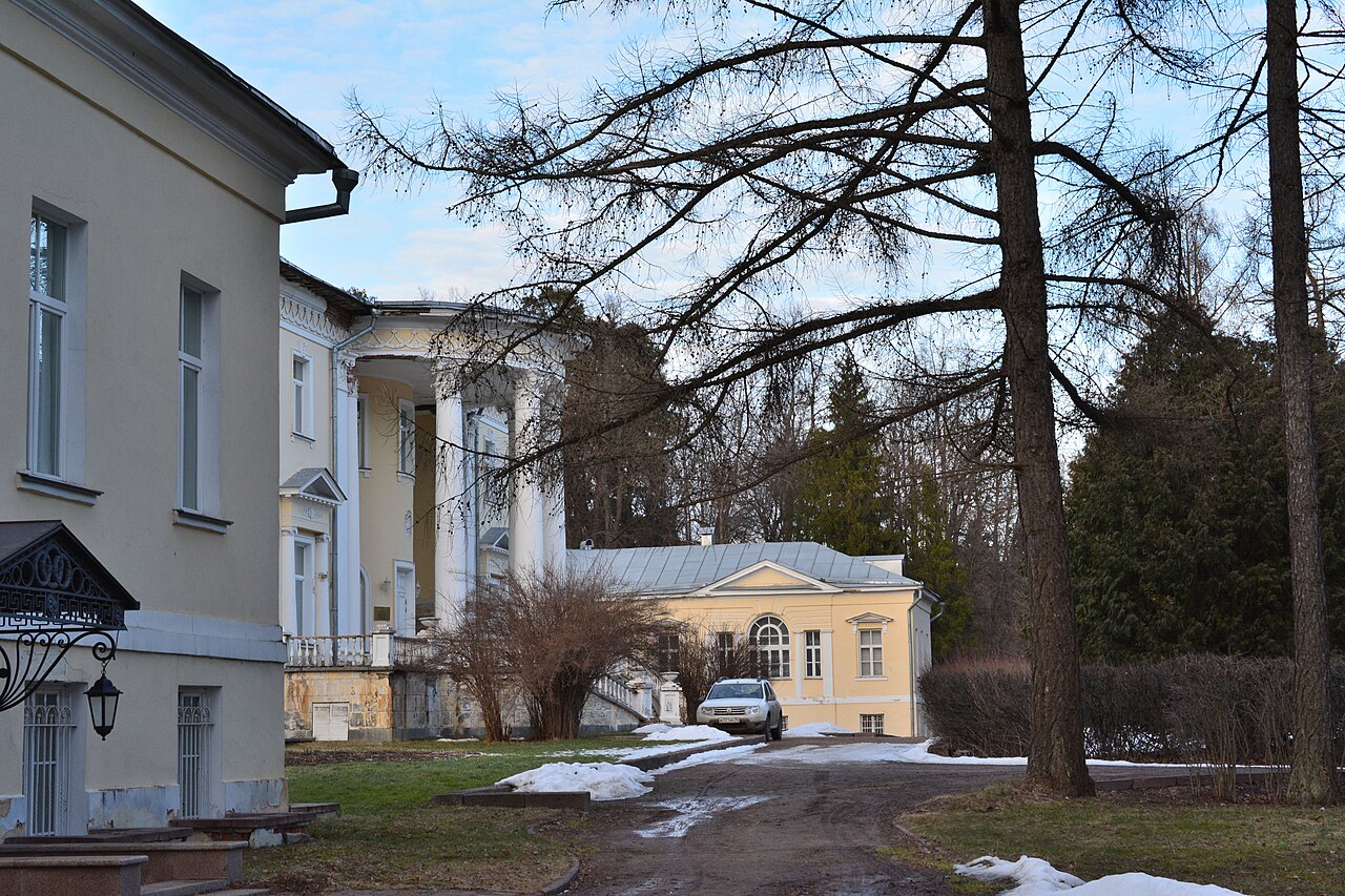 Усадебные постройки с колоннами и жёлтые корпуса среди деревьев, на земле лежит тающий снег.